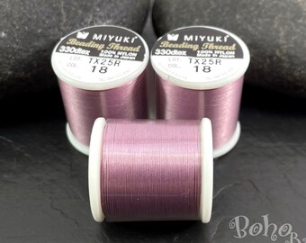 Op de afbeelding: Drie spoelen Miyuki Beading Thread in kleur 18, een lichtpaarse met een glinsterende afwerking. De spoelen zijn gelabeld "330dtex 100% Nylon Gemaakt in Japan" en "TX25R 18".