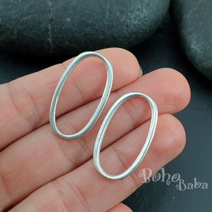 Könnte beinhalten: Zwei silberne ovale Sprungringe, etwa 2,5 cm im Durchmesser, werden in einer Hand gehalten. Die Ringe sind glatt und haben eine leicht strukturierte Oberfläche.