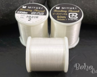 Può includere: Tre bobine di filo per perline bianco. Le bobine sono etichettate "MIYUKI Beading Thread" e hanno dimensioni e metraggi diversi. Le bobine sono su una superficie grigio scuro.