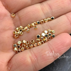 Puede incluir: Primer plano de una mano sosteniendo un montón de pequeñas cuentas doradas en forma de cubo. Las cuentas están esparcidas en la palma de la mano.