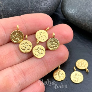 以下が含まれることがあります： オームのシンボルが刻まれた、金色の金属製のチャーム8個のクローズアップ。チャームは丸型で、上部に小さなループが付いており、ジュエリーやその他のクラフトに取り付けることができます。