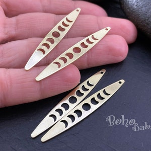 Può includere: Quattro pendenti allungati color ottone con un design a mezzaluna. Ogni pendente ha una serie di ritagli a mezzaluna lungo la sua lunghezza, con un piccolo foro all'estremità appuntita. I pendenti sono disposti su una superficie scura.