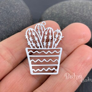 May include: A silver-colored metal charm in the shape of a potted cactus. The cactus has star-shaped cutouts, and the pot features a wavy design. The charm is held in a hand.