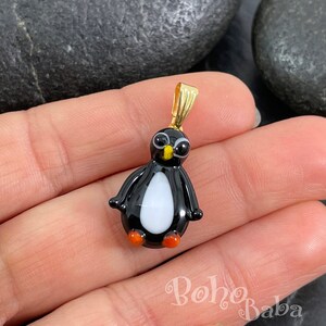 以下が含まれることがあります： 黒と白のガラス製のペンギンチャーム。オレンジ色の足と金色のベイルが付いています。チャームの高さは約2.5cmです。