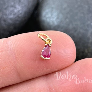 Op de afbeelding: Gouden bedel met een traanvormige rode edelsteen. De bedel heeft een lus aan de bovenkant om aan een ketting of armband te bevestigen.