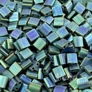 Puede incluir: Una imagen de primer plano de un montón de cuentas de semillas cuadradas de color verde azulado iridiscente. Las cuentas tienen un brillo metálico y son perfectas para la elaboración de joyas.