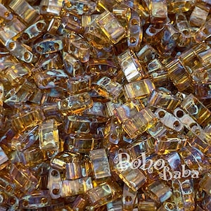 Op de afbeelding: Een close-up van een stapel kleine, rechthoekige, amberkleurige glazen kralen met iriserende vlekken. De kralen hebben twee gaten aan elke kant.