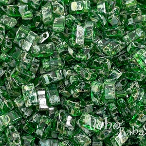 Puede incluir: Primer plano de una gran cantidad de pequeñas cuentas de vidrio rectangulares verdes con un brillo metálico. Las cuentas tienen una forma ligeramente irregular y están dispersas aleatoriamente.