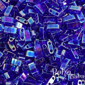 Op de afbeelding: Een close-up afbeelding van een stapel kleine, iriserende blauwe glazen kralen. De kralen zijn rechthoekig van vorm met twee gaten in elke kraal. De kralen zijn in een willekeurig patroon gerangschikt.