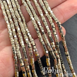 May include: Strands of small, metallic gold-colored square beads. The beads are strung together, creating multiple lines of the same design. The beads have a reflective surface, and the image is well-lit, highlighting the details of the beads.