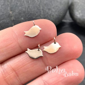 May include: Three small, shiny, bird-shaped charms with a loop at the top for hanging. The charms are a light gold colour and have a smooth, reflective surface. The charms are likely for jewellery making or crafting.