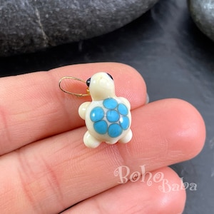 Può includere: Un piccolo ciondolo a forma di tartaruga in ceramica bianca con un guscio blu e bianco e occhi neri. La tartaruga ha un anello dorato per appenderlo.