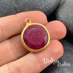 May include: A round pendant featuring a faceted, deep magenta-colored stone set within a textured, matte gold-toned metal bezel. The pendant includes a small circular loop at the top for attachment, showcasing a vibrant, polished gemstone surface.
