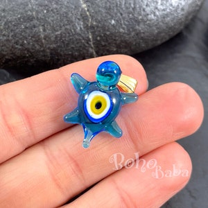 以下が含まれることがあります： 金色の輪と青い邪眼のデザインが施された青いガラス製の亀のチャーム。このチャームは幸運と保護の象徴です。