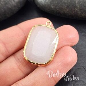 May include: A white gemstone pendant with a gold-toned frame. The pendant has a rectangular shape with a slightly rounded edge. The pendant is on a white background.