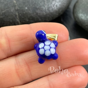 Può includere: Un piccolo ciondolo a forma di tartaruga in vetro blu e bianco con un'anellina dorata. La tartaruga ha un guscio dettagliato con macchie bianche.