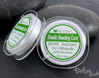 Puede incluir: Dos carretes de cordón elástico para cuentas de color blanco, cada uno en un recipiente de plástico transparente con una etiqueta verde y blanca. La etiqueta dice "MIYUKI Elastic Beading Cord H3198E 5m(5.5yds) 0.6~0.8mm dia 100% Polyurethane Made in Japan."