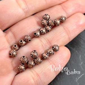 Peut inclure: Une collection de petites perles métalliques rondes de couleur cuivre, présentant un motif texturé en relief avec un point central et des lignes rayonnantes. Les perles sont disposées en deux rangées lâches, révélant leur fini métallique.
