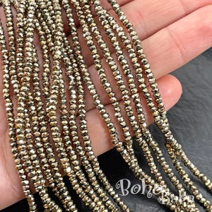 Peut inclure: Plusieurs brins de petites perles dorées à facettes. Les perles sont enfilées ensemble, créant plusieurs lignes du même matériau. Les perles ont un éclat métallique et sont tenues dans une main. Ces perles sont probablement destinées à la fabrication de bijoux.