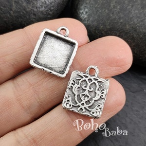 Puede incluir: Dos colgantes de metal de tono plateado. Un colgante es un cuadrado con un diseño de filigrana decorativo. El otro colgante es un cuadrado con una superficie lisa.