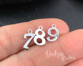 May include: Three silver metal number charms, 7, 8, and 9, with jump rings for attaching to jewelry or other crafts.  The charms are on a hand.