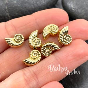 Pode incluir: São exibidos seis pequenos pingentes dourados em forma de concha. Cada pingente é uma réplica detalhada de uma concha de nautilus, com um design em espiral. Os pingentes são dispostos sobre um fundo neutro, mostrando seu brilho metálico. O tema geral é a fabricação de joias ou artesanato.