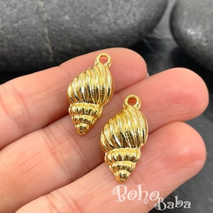 Könnte beinhalten: Zwei goldfarbene Muschelanhänger mit strukturierter Oberfläche und einer Schlaufe zum Befestigen an Schmuck. Die Anhänger werden in einer Hand gehalten. Der Text "Boho Baba" ist im Bild zu sehen.