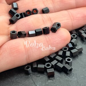 May include: A collection of small, black, hexagonal beads with a hole through the center. The beads are scattered across a hand, with the text "Boho Baba" visible in the lower left corner. These beads are ideal for jewelry making.