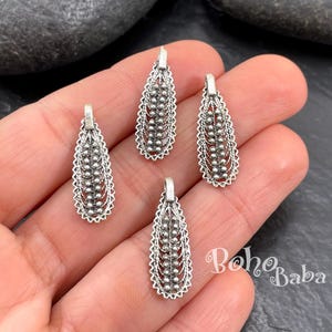 A hand holding four elongated antique silver-plated filigree teardrop charms with intricate cut-out wirework, granulated dotted details, and a solid top loop.