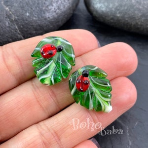 Può includere: Un paio di orecchini in vetro verde e bianco con una coccinella su ogni foglia. Gli orecchini sono piccoli e delicati.
