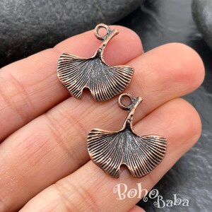 A close-up photograph displays two antique copper plated ginkgo leaf charms resting on a person's fingers against a dark rock background.