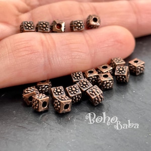 May include: Close-up of small, cube-shaped beads with a bronze finish. Each bead features a detailed, textured pattern of small dots. The beads are scattered on a dark surface, with some held in a hand. The text "Boho Baba" is visible.