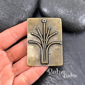 Puede incluir: Un colgante rectangular de color latón antiguo con un diseño de árbol estilizado. El colgante tiene una superficie texturizada y está sujeto en una mano. El texto "Boho Baba" es visible en la esquina inferior derecha.