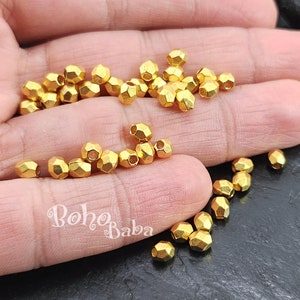 Puede incluir: Primer plano de una mano sosteniendo un montón de cuentas de metal de color dorado. Las cuentas son facetadas y tienen una forma ligeramente irregular. Las cuentas están dispersas sobre una superficie oscura.