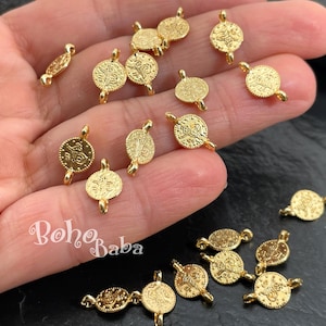 以下が含まれることがあります： 金色の金属製ジュエリーパーツのクローズアップ。エンボス加工が施されたコインのような丸いチャームと楕円形のコネクタが見えます。チャームには、他のジュエリーパーツに取り付けるための小さなループが付いています。