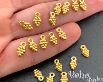 May include: A collection of small, gold-colored metal charms shaped like clusters of grapes. Each charm features a textured, beaded surface and a small loop at the top for attachment. The charms are displayed against a dark background.