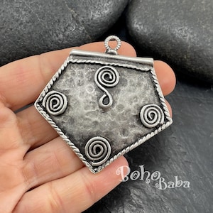 May include: A silver-toned pendant with a geometric shape and spiral designs. The pendant has a textured surface and a decorative rope-like border. The design includes three spiral accents and a central S-shaped swirl. A small loop is at the top for hanging.