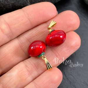 Peut inclure: Deux pendentifs en verre rouge avec une bélière dorée et une bordure verte. Les pendentifs ont la forme d'une tomate ou d'un cœur.