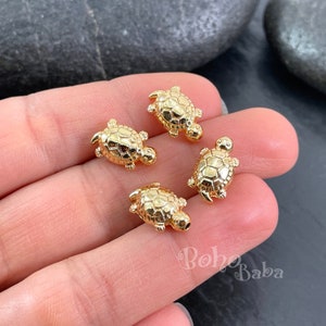 May include: Four gold-coloured turtle charms, each with a detailed shell design, are displayed on a hand. The charms are small and perfect for jewellery making or crafting.