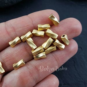 May include: A close-up of a hand holding a group of gold-colored metal beads. The beads are twisted and have a shiny finish. The beads are approximately 1/2 inch in length.