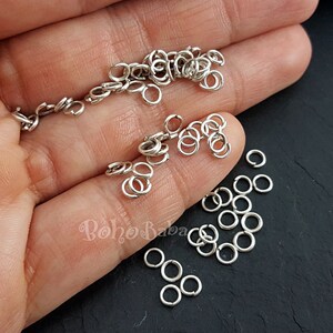 May include: A close-up of a hand holding a large quantity of silver jump rings. The jump rings are small, circular, and have a smooth, polished finish.