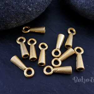 May include: A group of 10 gold-toned metal cord end caps with a loop at the top. The cord end caps are approximately 2.5 cm long and have a tapered shape.