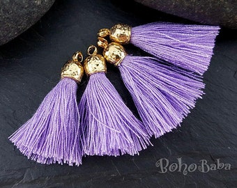 May include: Four lavender tassels with gold-colored caps. The tassels are made of fine threads, and the caps have a hammered texture. The tassels are arranged on a dark, textured surface, with a large, dark stone in the background.