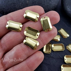 May include: A hand holding a variety of gold-colored metal rectangular embellishments with two holes in each. The embellishments are scattered on a dark gray surface.
