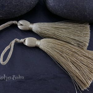May include: Two beige tassels with long, thin strands. The tassels are tied together with a thin cord.