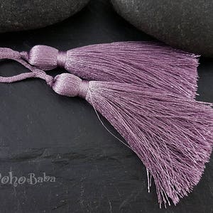Könnte beinhalten: Zwei violette Quastenbesätze mit einer langen Franse. Die Quasten bestehen aus weichen, seidenartigen Fasern und sind perfekt, um jedem Projekt einen Hauch von Eleganz zu verleihen.