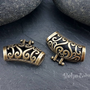 Op de afbeelding: Twee sierlijke, antiek messingkleurige metalen sieradenonderdelen. Elk stuk heeft een donker interieur met wervelende, opengewerkte ontwerpen. De onderdelen hebben een gebogen vorm en kleine, rechthoekige bevestigingspunten.