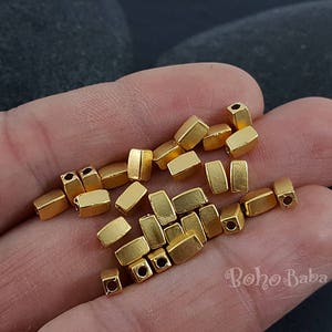 May include: A pile of gold-coloured metal beads, each with a rectangular shape and a small hole in the centre. The beads are arranged in a random pattern on a hand.
