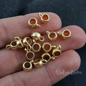 Puede incluir: Primer plano de una colección de anillas de salto de metal dorado. Las anillas son todas del mismo tamaño y forma, y están dispuestas en una pila suelta sobre una mano.