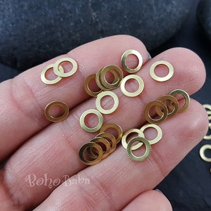 Puede incluir: Primer plano de una mano sosteniendo un montón de pequeños anillos de metal dorados. Los anillos son todos del mismo tamaño y forma, y están dispuestos en un patrón aleatorio. También hay algunos anillos dispersos sobre una superficie oscura.